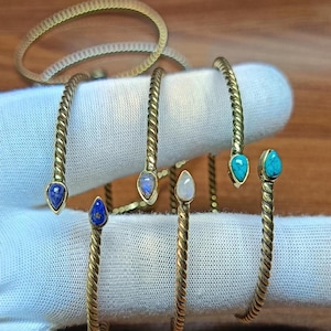 May include: Gold-toned metal bracelets with a twisted design. Some bracelets feature teardrop-shaped gemstones in blue, turquoise, and white. The bracelets are displayed on a white surface.