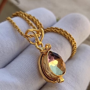 May include: A gold-colored necklace with a teardrop-shaped pendant. The pendant features a faceted, multi-colored gemstone with hues of purple, yellow, and green. The chain is a delicate, woven design. The necklace is displayed against a white background.