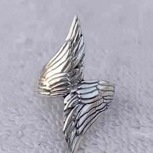 May include: A silver-colored ring with a wing design. The ring features two detailed wings that wrap around the finger. The wings have textured feather details, creating a realistic appearance. The ring is likely made of metal and has a polished finish.
