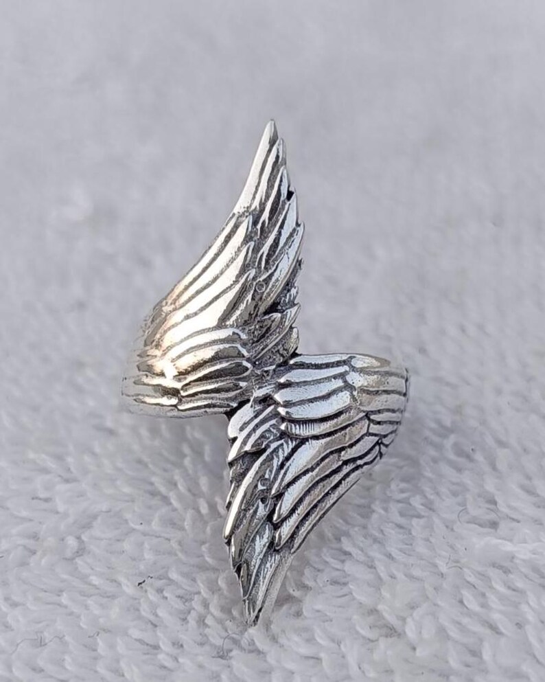 May include: A silver-colored ring with a wing design. The ring features two detailed wings that wrap around the finger. The wings have textured feather details, creating a realistic appearance. The ring is likely made of metal and has a polished finish.
