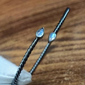 May include: An open cuff bracelet featuring a dark, oxidised metal band with a twisted rope texture. The two ends of the bracelet are capped with small, pear-shaped moonstones that exhibit a soft, iridescent blue glow against the dark metal.