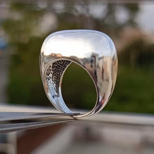 May include: A polished silver ring with a rounded, rectangular top and a textured interior band. The ring is held by tweezers, highlighting its reflective surface and unique design. The ring's design is modern and minimalist.