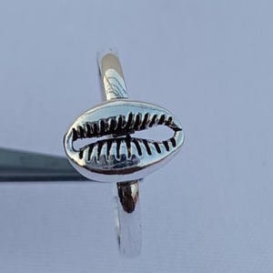 Peut inclure: Une bague en argent ornée d'un motif de coquillage cauri détaillé. La bague a une finition polie et une bande simple. Le coquillage cauri est le point central, avec des textures et des courbes complexes. La bague est posée sur un fond clair.