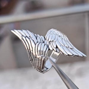 May include: A silver-colored ring featuring a detailed wing design. The wings are spread out, with intricate feather detailing. The ring is shown close-up, highlighting the metal's texture and the craftsmanship of the jewelry.
