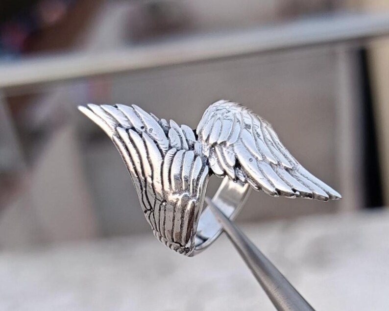 May include: A silver-colored ring featuring a detailed wing design. The wings are spread out, with intricate feather detailing. The ring is shown close-up, highlighting the metal's texture and the craftsmanship of the jewelry.