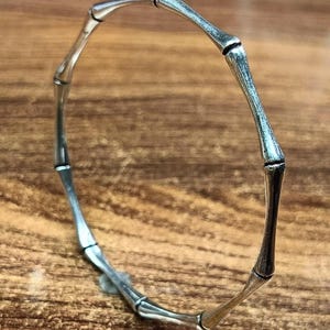 May include: A silver-toned bangle bracelet featuring a segmented design that mimics the appearance of bamboo stalks. The metal surface has a polished finish with darkened recessed areas at the joints to emphasize the individual segments.