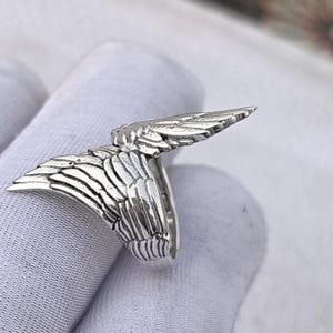May include: A silver-colored ring featuring a detailed wing design. The ring's wings are spread out, showcasing intricate feather detailing. The ring is likely made of sterling silver or a similar metal, and is designed to be worn on a finger.