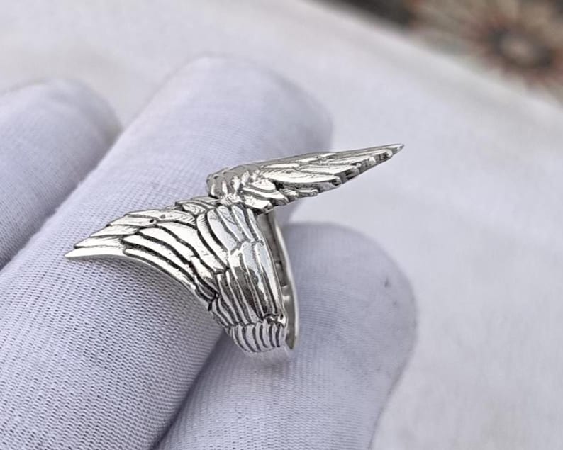 May include: A silver-colored ring featuring a detailed wing design. The ring's wings are spread out, showcasing intricate feather detailing. The ring is likely made of sterling silver or a similar metal, and is designed to be worn on a finger.