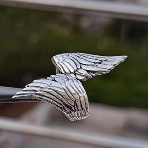 May include: A silver-colored ring with a detailed wing design. The ring features two wings, each with intricate feather detailing, creating a symmetrical and elegant appearance. The ring is likely made of metal and is designed to be worn on a finger.