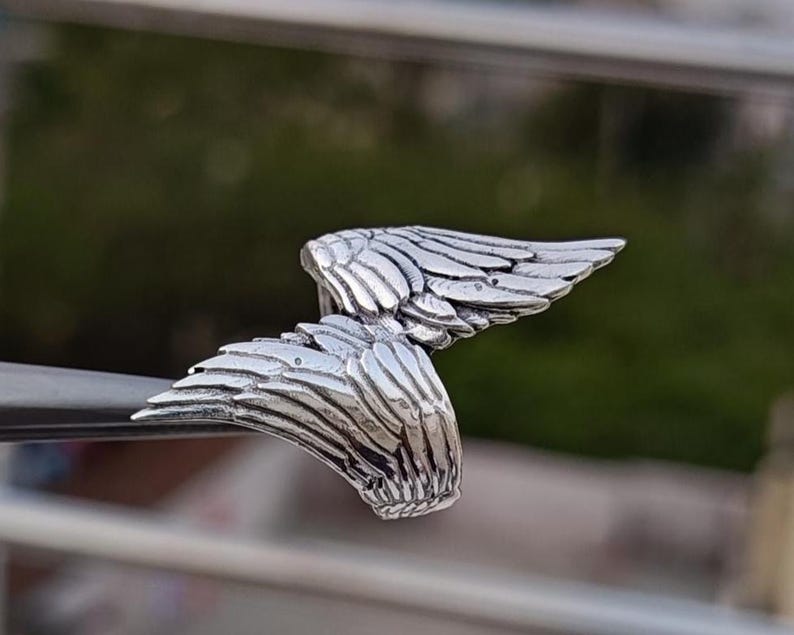 May include: A silver-colored ring with a detailed wing design. The ring features two wings, each with intricate feather detailing, creating a symmetrical and elegant appearance. The ring is likely made of metal and is designed to be worn on a finger.