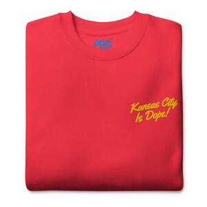 Puede incluir: Una sudadera roja con el texto "Kansas City Is Dope!" bordado en escritura amarilla. La sudadera también presenta el texto "Midwest is Dope" en escritura azul cerca del cuello.