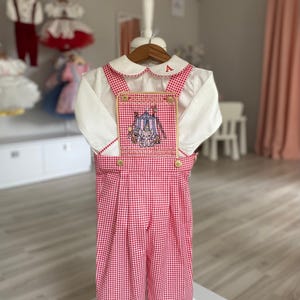 Könnte beinhalten: Ein weißes Hemd mit einer rot-weiß karierten Latzhose. Die Latzhose hat einen aufgestickten Aufnäher mit Zirkusmotiv. Das Hemd hat einen weißen Kragen mit roter Paspelierung und dem Buchstaben "A" auf dem Kragen. Das Outfit wird auf einem Kleiderbügel präsentiert.
