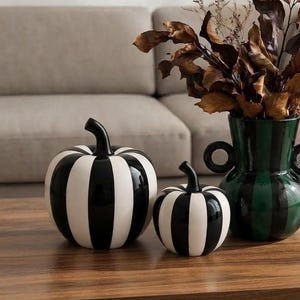 May include: Two ceramic pumpkins with vertical black and white stripes sit beside a dark green and black striped vase with two circular handles. The vase contains a cluster of dried brown leaves and branches, creating a decorative arrangement.
