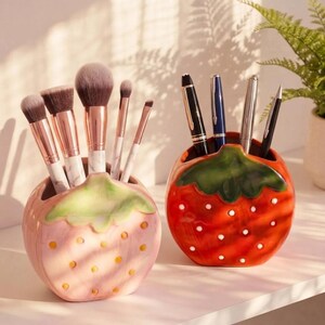 May include: Two ceramic strawberry-shaped holders. The left holder is light pink with yellow seeds and holds five makeup brushes with rose gold ferrules. The right holder is red with white seeds and holds four pens with metallic clips.
