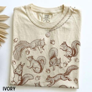 Könnte beinhalten: Ein elfenbeinfarbenes T-Shirt mit Rundhalsausschnitt und einem braunen Strichzeichnungsmuster aus mehreren Eichhörnchen und Eicheln, die über den Stoff verteilt sind. Das Innenetikett zeigt den Text Comfort Colors und eine silberne Kette liegt darauf.