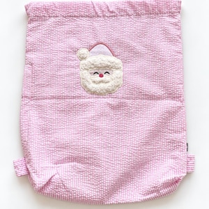 May include: A light pink and white seersucker drawstring backpack featuring an embroidered applique of a Santa Claus face with a textured white beard, a pink hat, and a small pink nose, centred on the front of the fabric bag.