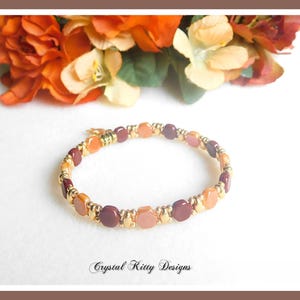 May include: A beaded bracelet featuring a repeating pattern of hexagonal beads in shades of orange, peach, and deep maroon, interspersed with small metallic gold spacer beads. The text Crystal Kitty Designs is printed at the bottom of the image.