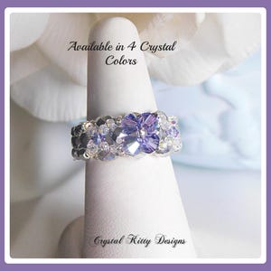 May include: A beaded ring featuring a central purple crystal flower surrounded by smaller clear and metallic grey crystals. The text Available in 4 Crystal Colors and Crystal Kitty Designs is printed on the display stand holding the ring.