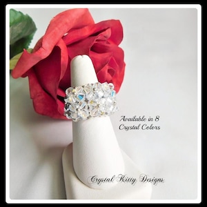 May include: A wide ring crafted from clear, faceted crystals arranged in a floral pattern. The text "Available in 8 Crystal Colors" and "Crystal Kitty Designs" is printed nearby. A large red rose is positioned behind the ring display.