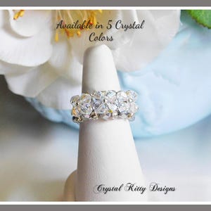 May include: A ring made of clear, faceted crystal beads arranged in a multi-row band. The text "Available in 5 Crystal Colors" and "Crystal Kitty Designs" is visible. The ring is displayed on a white conical stand against a floral background.