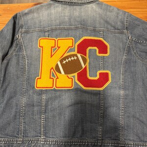 May include: A denim jacket with a yellow "K" and a red "C" patch on the back. A brown football is centered within the "KC" design. The jacket is a light blue wash.