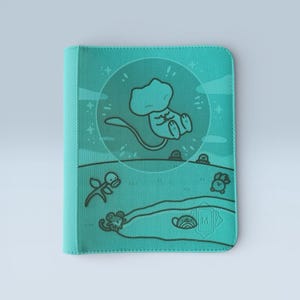 May include: Teal card binder with a cartoon illustration. The design features a floating creature inside a bubble, with other cartoon characters and a landscape. The binder has a textured appearance and a clean, minimalist design.
