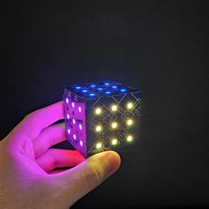 May include: A black cube with a geometric design and illuminated dots in blue, yellow, and purple. The cube is held in a hand against a dark background, showcasing its glowing features.