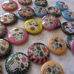 May include: A collection of colorful round buttons featuring a sugar skull design. The buttons have black, white, pink, blue, yellow, and orange colors. The sugar skulls have intricate details and are surrounded by floral patterns.