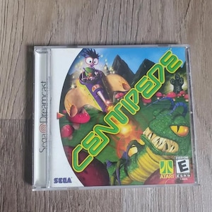 May include: A Sega Dreamcast video game case for Centipede featuring a green creature with sharp teeth, a character in a purple and yellow vehicle, and the title Centipede in large green letters against a colorful, illustrated background.
