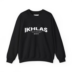 Op de afbeelding: Zwarte crewneck sweatshirt met het woord "IKHLAS" in grote witte hoofdletters op de borst. Daaronder staat een kleinere tekst: "Oprecht zuivert de intentie, kleine daden veranderen in bergen van beloning."