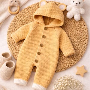 以下が含まれることがあります： クマの耳が付いたフード、木製のボタン、白い袖口が付いた黄色のニットベビーロンパース。画像には、ウサギの形をした木製歯固め、白いテディベア、お揃いのブーティーが含まれています。ロンパースは織りマットの上にあります。