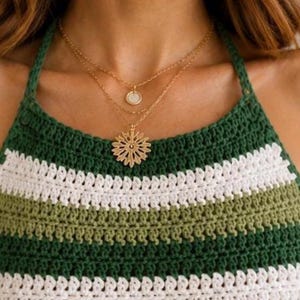 Pode incluir: Um colar dourado em camadas com um pequeno pingente e um pingente maior e intrincado. O colar &eacute; usado sobre um top halter de croch&ecirc; listrado verde, branco e verde-oliva.