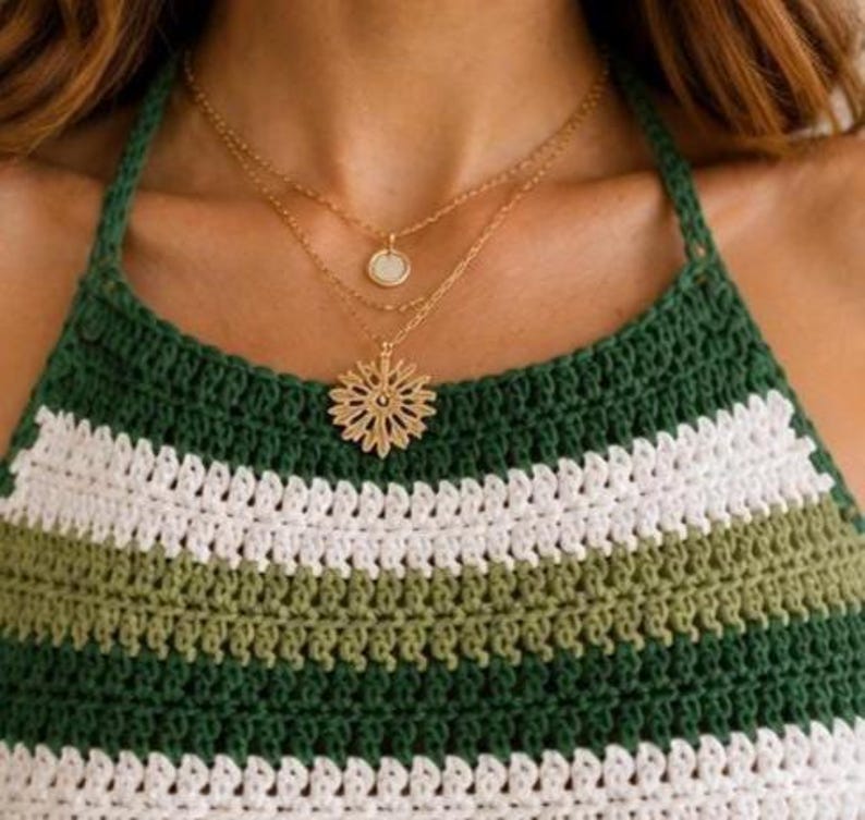 Pode incluir: Um colar dourado em camadas com um pequeno pingente e um pingente maior e intrincado. O colar &eacute; usado sobre um top halter de croch&ecirc; listrado verde, branco e verde-oliva.