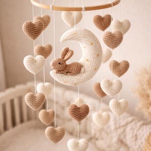 May include: A handmade baby mobile featuring a wooden hoop, crocheted hearts in cream and brown, and a cream-colored moon with a small brown bunny. The mobile is suspended by white strings, creating a whimsical nursery decoration.