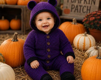 May include: A purple crocheted long-sleeve romper with a hood featuring two round ears. The garment has a black trim along the hood and cuffs, and a vertical row of brown buttons down the front. The texture is consistent throughout the piece.