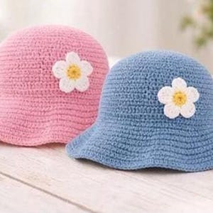Puede incluir: Dos sombreros de sol de ganchillo, uno rosa y otro azul. Cada sombrero tiene una flor blanca con centro amarillo. Los sombreros tienen ala ancha y probablemente son para niños.