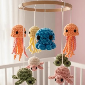 Könnte beinhalten: Handgefertigtes Babymobile mit gehäkelten Meeresbewohnern. Das Mobile umfasst orangefarbene Quallen, einen blauen Oktopus, gelbe Quallen sowie grüne und rosa Schildkröten. Die Kreaturen hängen an einem Holzring mit weißen Schnüren.