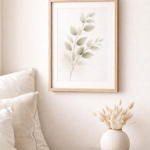 Könnte beinhalten: Ein gerahmter Aquarelldruck eines belaubten Zweigs in sanften Grün- und Grautönen. Das Kunstwerk wird an einer weißen Wand über einem Nachttisch aus Holz mit einer weißen Vase mit getrockneten Blumen ausgestellt. Beige Kissen sind links.