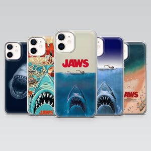 May include: Five phone cases with designs from the film "Jaws." The cases display images of sharks, the film's title, and beach scenes. The cases are transparent, with the designs printed on them. The cases are designed to fit a smartphone.