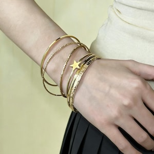 May include: A selection of gold bangles on a wrist. The bangles feature smooth, textured, and star-shaped designs. The star-shaped bangle is a key element, adding a playful touch. The bracelets are worn on a wrist.