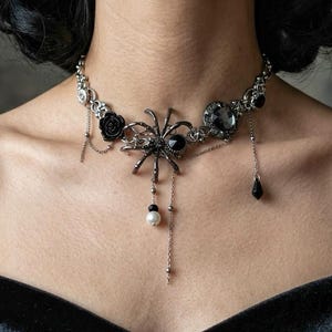 May include: A silver-toned choker necklace featuring a central spider charm, a black rose accent, and a round faceted black stone. The piece includes delicate silver chains, a single white bead, and a black teardrop pendant hanging from the structure.