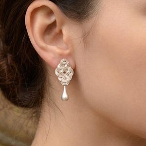 May include: Silver earrings with a woven knot design and a teardrop pendant. The earrings are a light silver colour and are worn on the ear. The earrings are a type of jewellery.