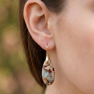 May include: A gold-colored earring with a teardrop-shaped stone. The stone features a mosaic of colors, including beige, red, and blue. The earring is hanging from a person's ear.