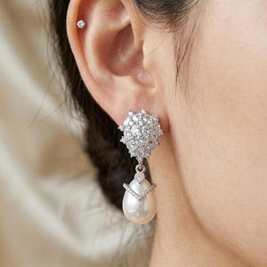 May include: A silver-toned drop earring featuring a cluster of clear, faceted stones at the top and a smooth, white teardrop-shaped pearl suspended below. A small, V-shaped silver accent set with tiny clear stones sits atop the pearl.