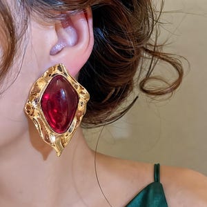May include: A large, statement earring featuring a central, smooth, deep red, diamond-shaped stone set within a textured, irregular, gold-toned metal frame. The metal surface has a hammered, organic appearance with undulating edges.