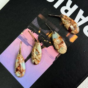 May include: Four teardrop-shaped earrings with a natural stone appearance. The earrings have a mix of beige, brown, and blue-green tones, with silver-colored hooks. The earrings are displayed on a black surface with a colorful background.