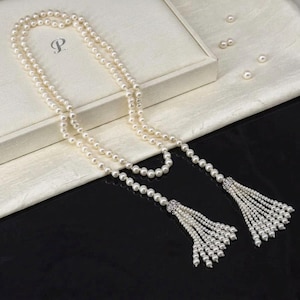 May include: A long pearl necklace with a tassel design. The necklace is made of multiple strands of small, round pearls. The tassels are decorated with smaller pearls and sparkling details. The necklace is presented on a white surface.