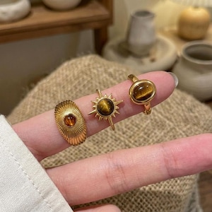 May include: Three gold rings, each featuring a brown gemstone. One ring has a sunburst design, another an oval stone, and the third a textured, oval face. The rings are displayed on a finger.