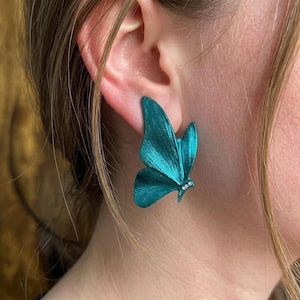 Könnte beinhalten: Ein teal-farbener, schmetterlingsförmiger Ohrring mit einer texturierten, metallischen Oberfläche und drei kleinen, klaren, runden Steinen, die nahe der Flügelbasis eingelassen sind. Der Ohrring befindet sich am Ohrläppchen und zeigt seine Form.