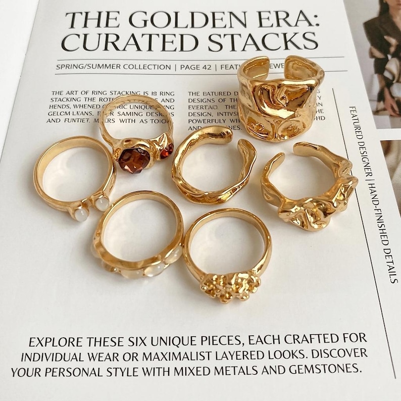May include: A collection of six gold-toned rings with unique designs, including textured, gemstone-adorned, and open-ended styles. The rings are displayed on a magazine page with the headline "THE GOLDEN ERA: CURATED STACKS."